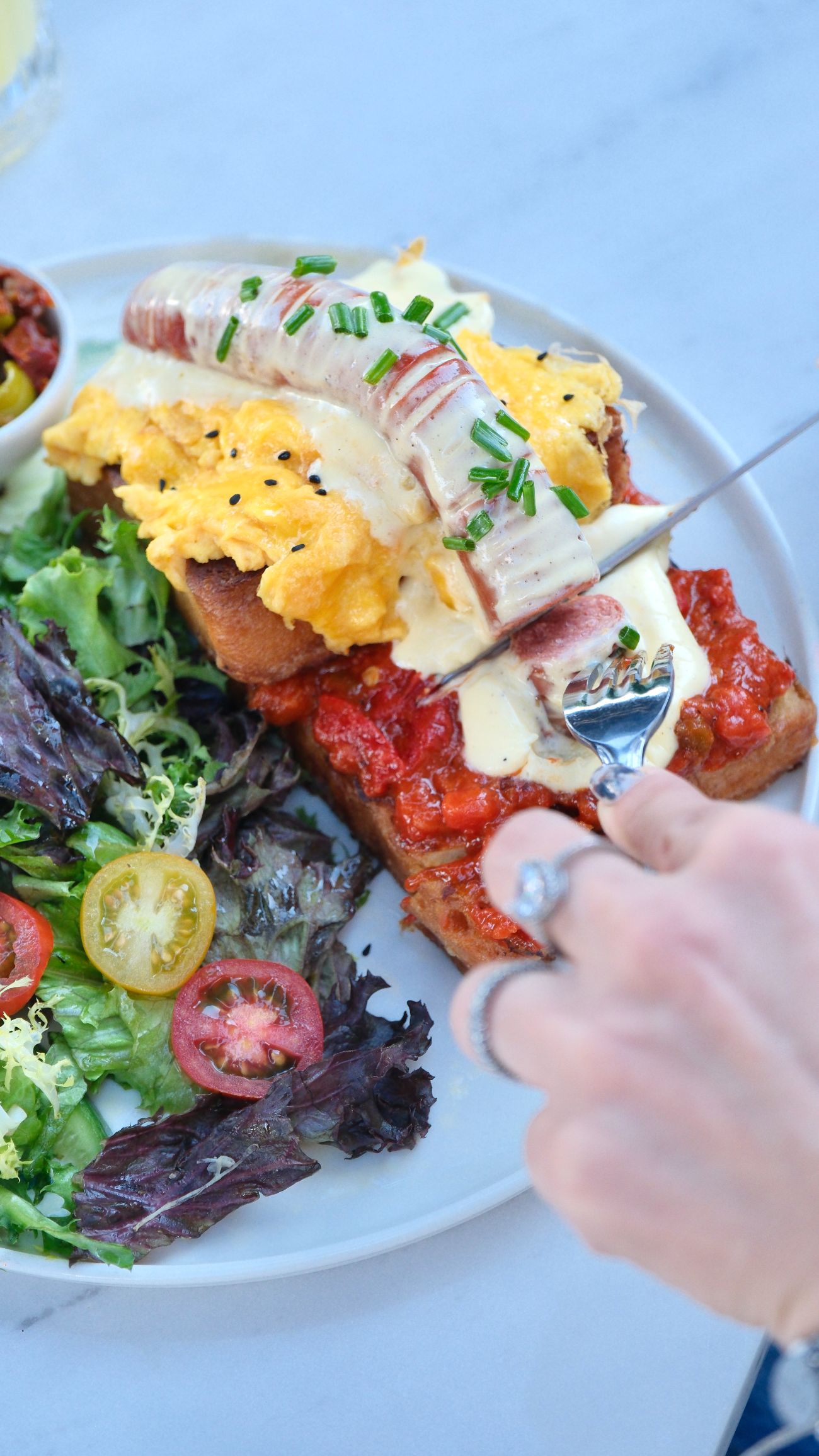 Sausage Benedict - Brioche bread, lutenitsa balkan sauce, scrambled eggs, beef frankfurter sausage, hollandaise sauce, chives, mescolonise salad and green olives.