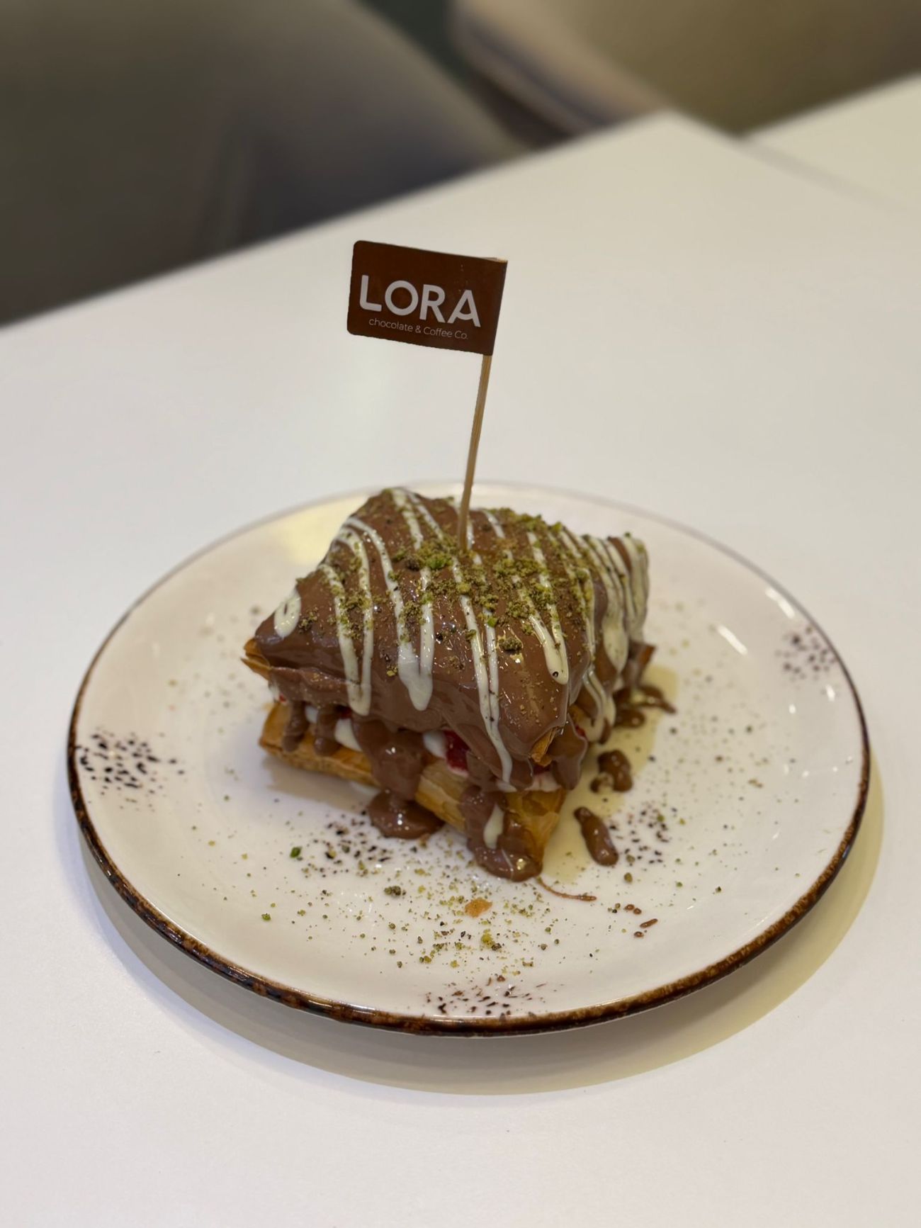 CROCCANTE (Yeni) - Pişirilmiş Milföy arasına özel Lora kreması, Çilek, Muz üzerinde toz antep fıstığı, sütlü ve beyaz çikolata ile servis edilir.